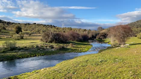 filming-with-a-camera-turn-of-a-stream-of-crystal-clear-waters-at-sunset-with-its-green-banks-and-shadows-projected-in-fields-of-trees-with-a-blue-sky-with-white-clouds-in-winter-in-Avila-Spain