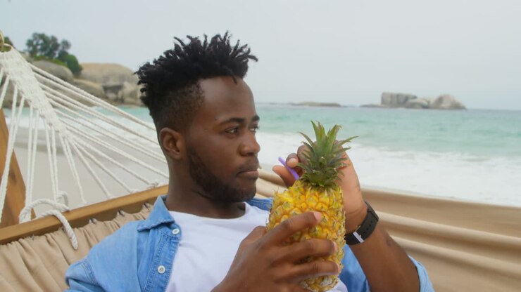 Vista frontale di uomo afroamericano che beve succo d'ananas sulla spiaggia 4k