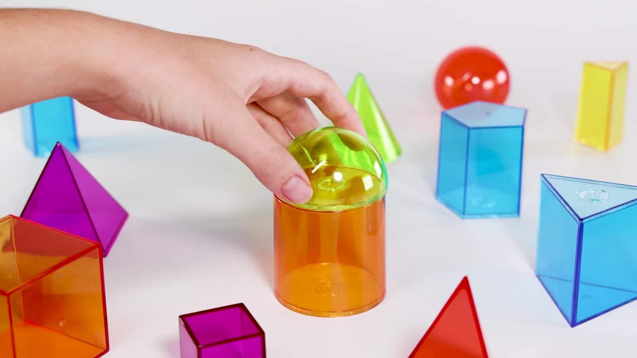 Una mano coloca un hemisferio verde transparente sobre un cilindro naranja entre formas geométricas coloridas