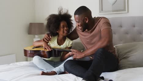 african american father and his daughter sitting on bed playing guitar together