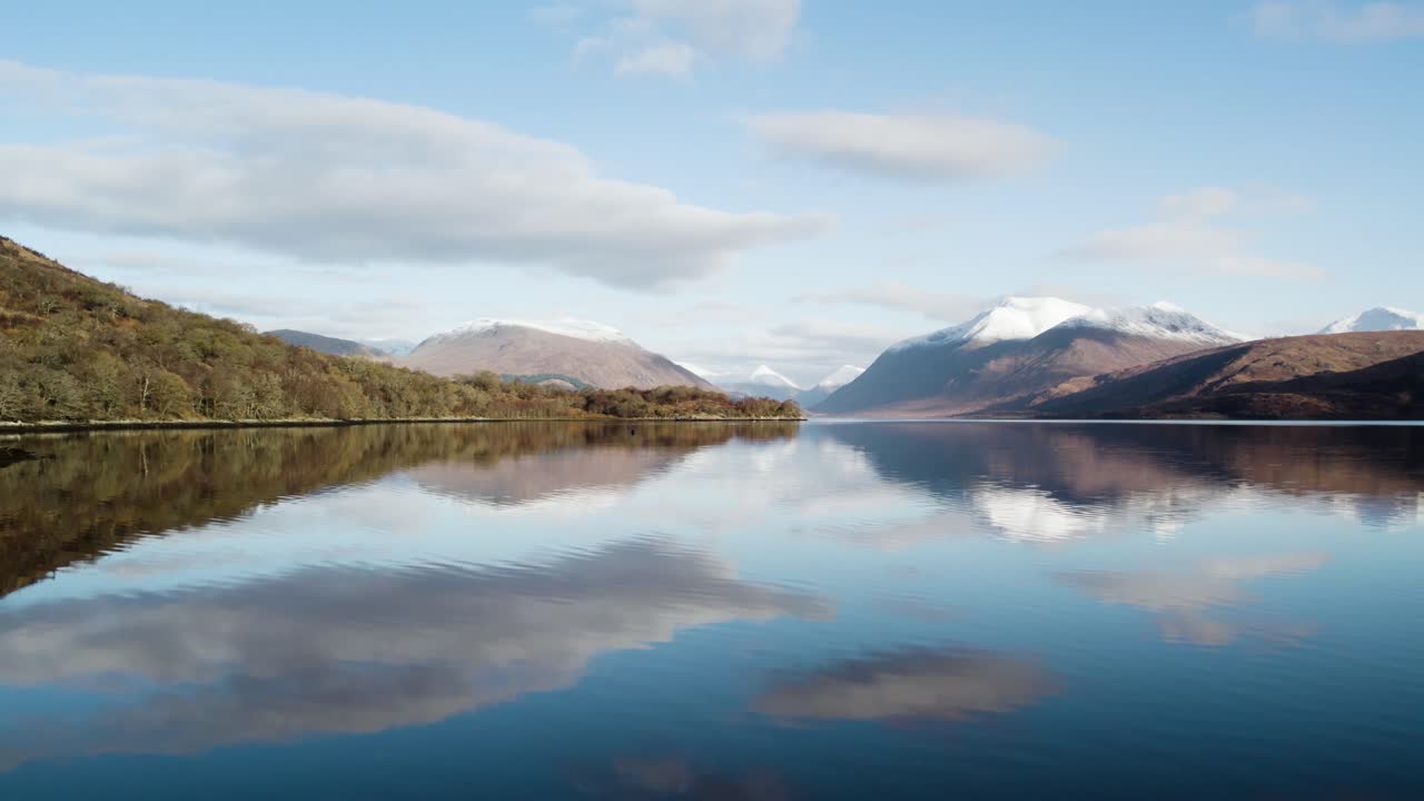 Aerial drone footage flying above the still, reflective water of Loch Etive revealing Glen Etive in the Scottish Highlands in winter with snow-capped mountains and native trees with blue sky and cloud