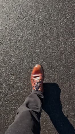 detailed first-person view of brown leather shoes walking across sunlit black asphalt, showing movement and texture.