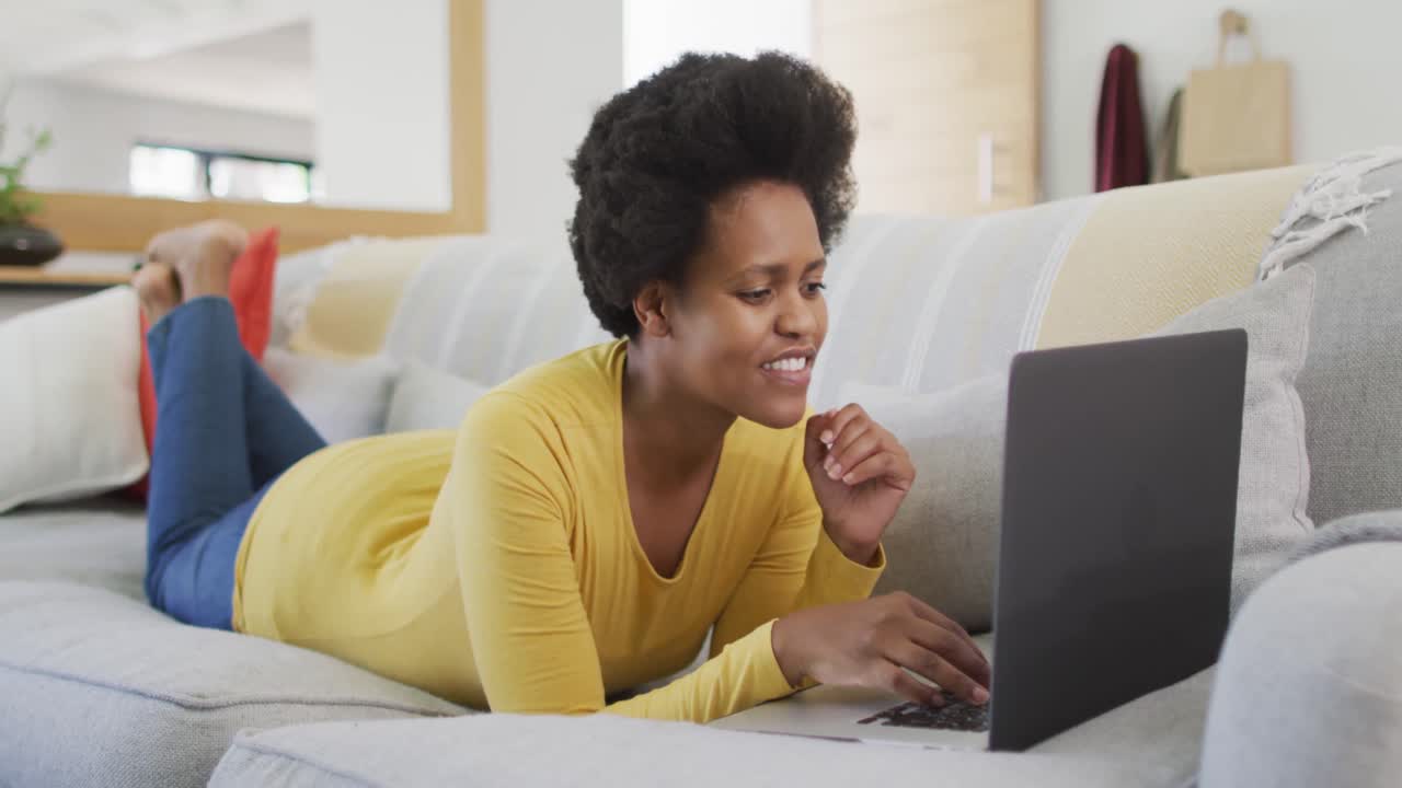 Happy african american woman laying on sofa using laptop