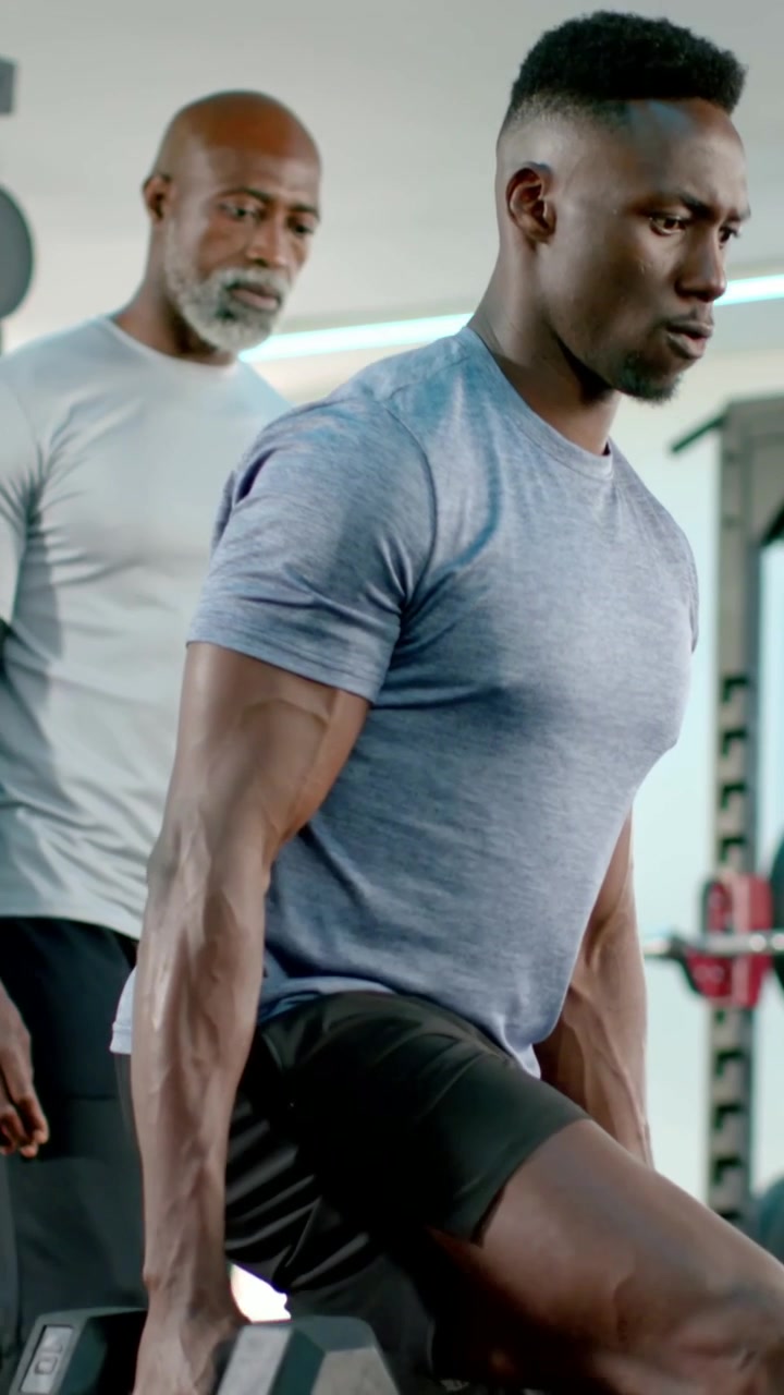 Vertical video: Holding hex weights, man doing lunges in gym for strength, in tee, coach watching