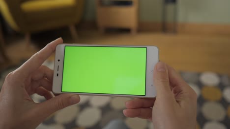 mujer joven en casa sentada en un sofá con un teléfono inteligente de pantalla verde en modo horizontal. niña usando teléfono móvil de pantalla táctil. niña utilizando teléfono inteligente, navegando por internet, viendo contenido de video, blogs. pov.