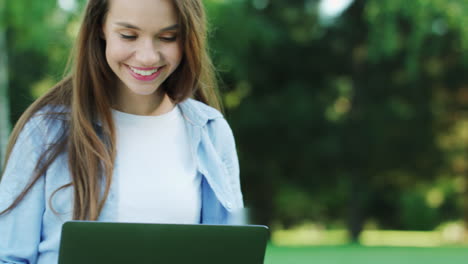 Smiling-woman-working-on-laptop-computer-in-city-park-at-summer-day