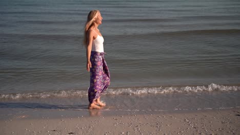 Cámara-Lenta-De-Mujer-Madura-En-Sarong-Caminando-En-Una-Playa-Y-Lanzando-Sus-Brazos-Al-Lado-Celebrando-La-Libertad,-Al-Amanecer-O-Al-Atardecer