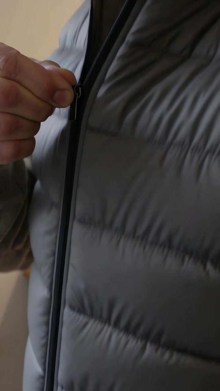 Vertical video: Aligning zipper, man wearing grey quilted vest pulling zipper up to collar at home