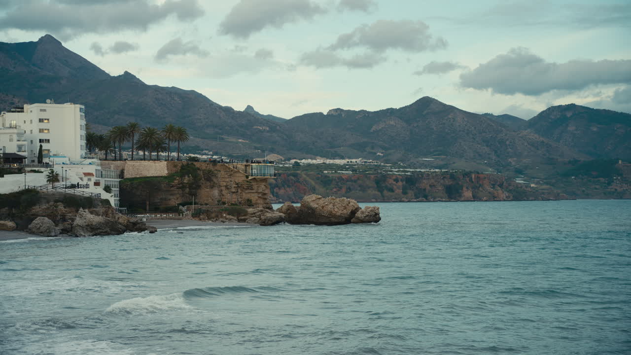 Wide coastal view of Balcon de Europa in Nerja, Spain, showing cliffside buildings, palm trees and rocky shoreline against the Mediterranean Sea