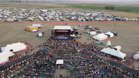 performers at a large outdoor music festival entertaining a large crowd in front of a stage and a campground with tents and recreational vehicles and mobile homes at the rear