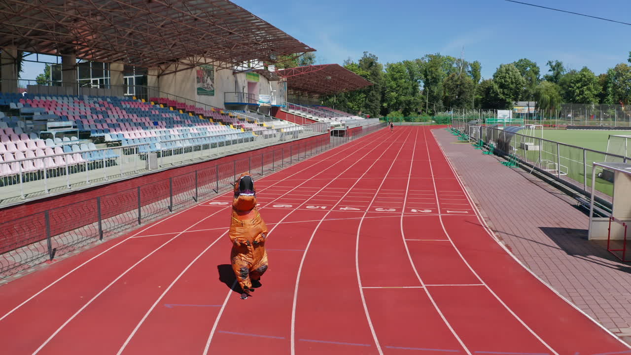 Animator wearing air inflatable costume of dinosaur walking along the stadium. Funny orange dinosaur costume on the red stadium track.