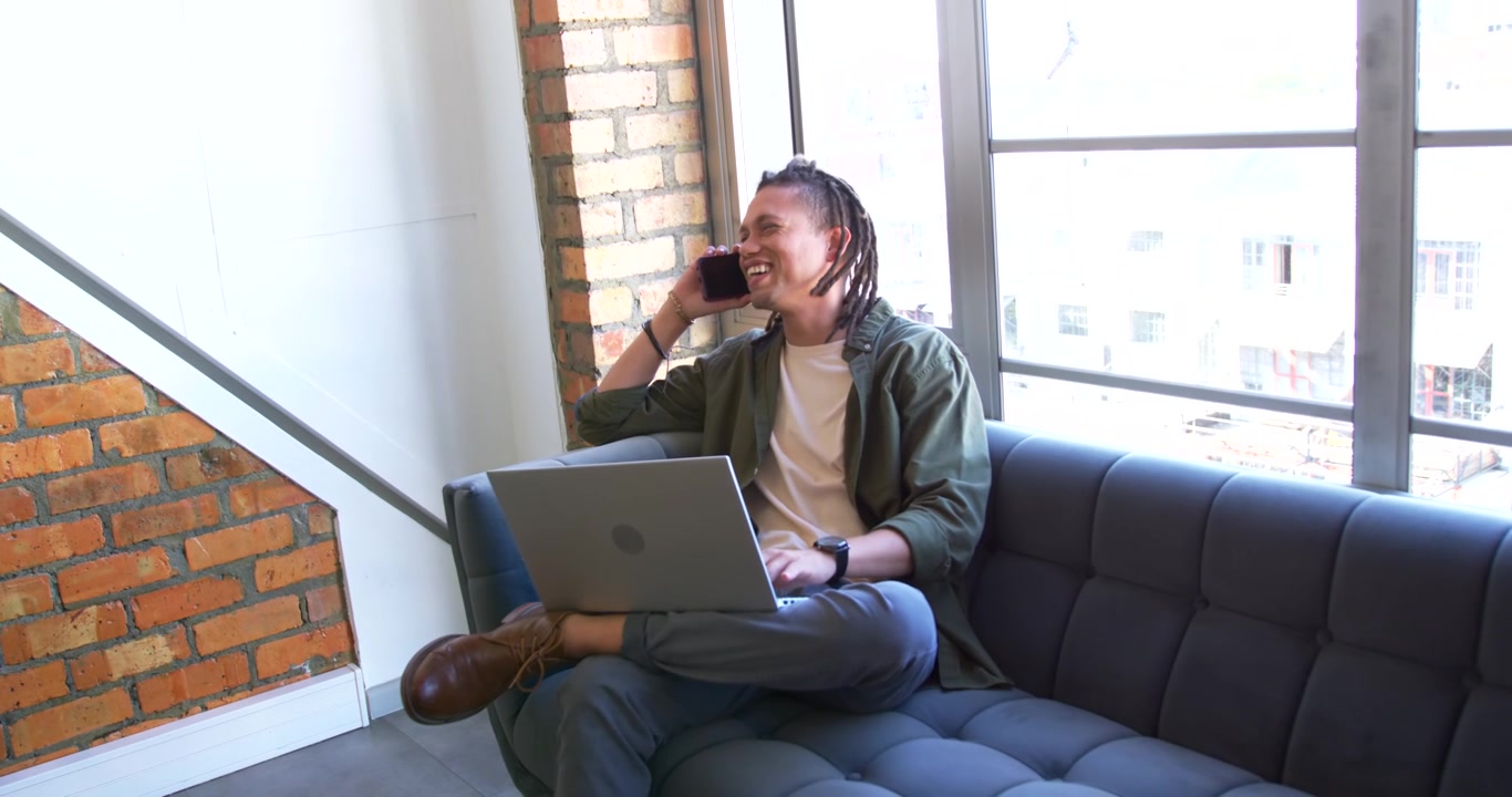African American man answering phone listening laughing typing on laptop on sofa by window for work