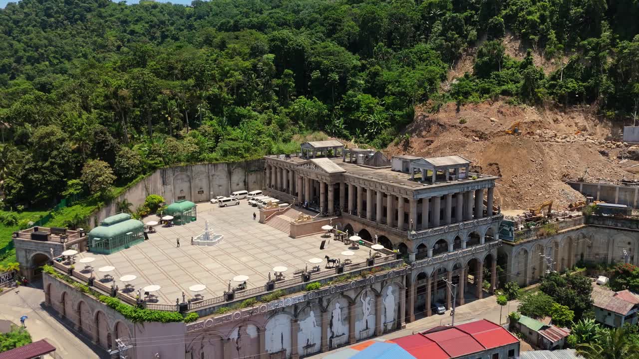Drone shot of a grand neoclassical-style building nestled on a hilltop, framed by tropical forest and ongoing hillside development