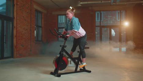 Mujer-Atlética-Montando-Una-Rutina-De-Entrenamiento-En-Bicicleta-Estacionaria-Giratoria-En-Un-Gimnasio-De-Niebla,-Pérdida-De-Peso-En-Interiores