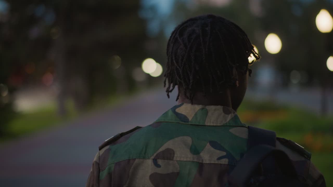 Black Soldier Walking Away Into Night Avenue, Rear View In Camouflage With Backpack, Street Bokeh And Lamppost Glow, Moving Forward From Service Toward New Life With Quiet Resolve