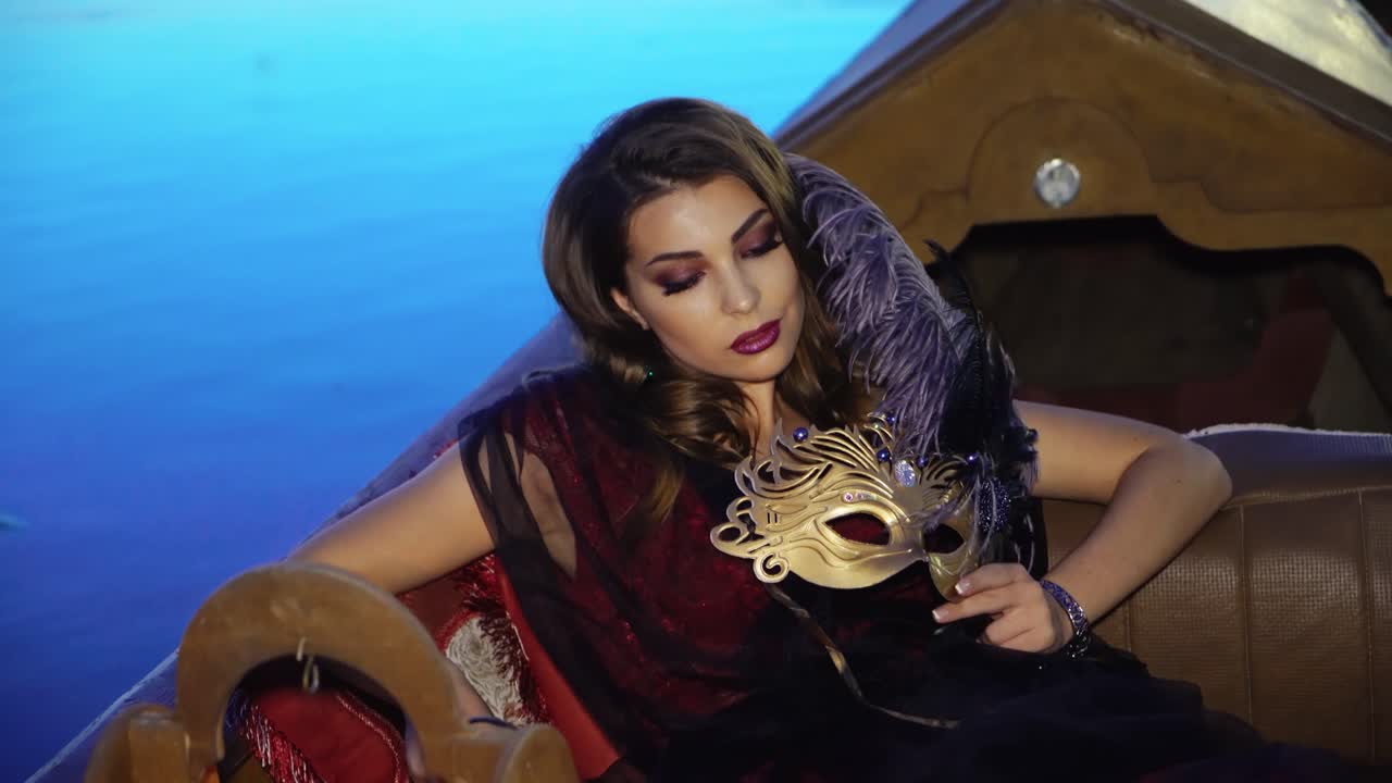 A gorgeous model in a dress is sitting in the gondola and holding a mask in golden colour with precious stones and a purple feather in her hand on the background of water in the evening.