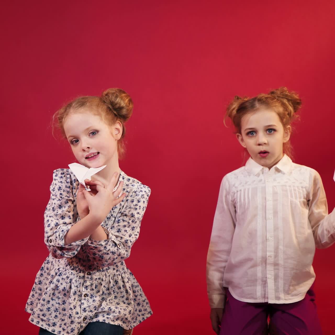 two little sisters with hairstyles are playing with paper origami in the form of planes on the background of red studio