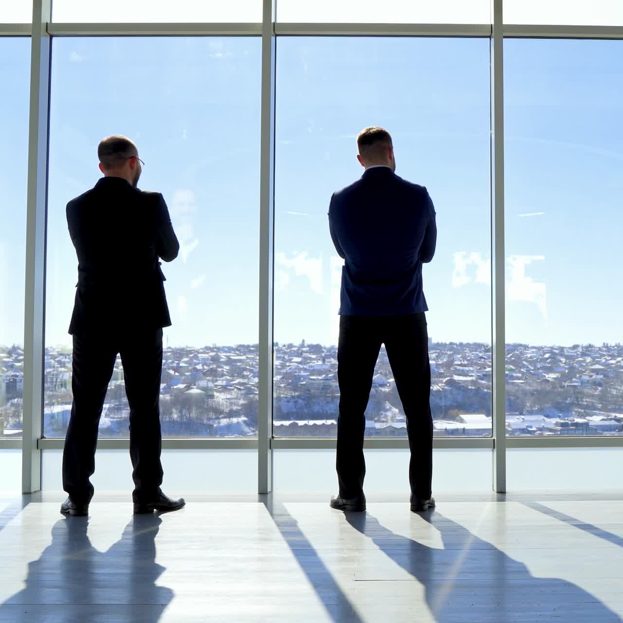 Reunión de emprendedores en la oficina junto a la ventana. Vista trasera de dos empresarios admirando el paisaje urbano a través de la ventana de la oficina y hablando sobre un nuevo proyecto