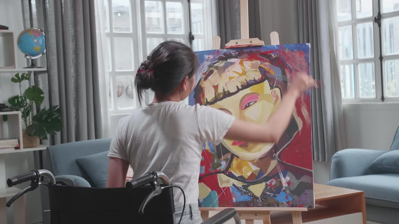 Back View Of Talented Female Artist Sitting On Wheelchair, Enthusiastically Using Paint Brush She Creates With Oil Painting. Creative Studio With Canvas