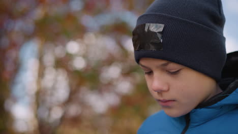 kid wearing black beanie with patch and blue jacket strolling with head bowed thoughtfully, surrounded by blurred vibrant autumn foliage and soft natural lighting