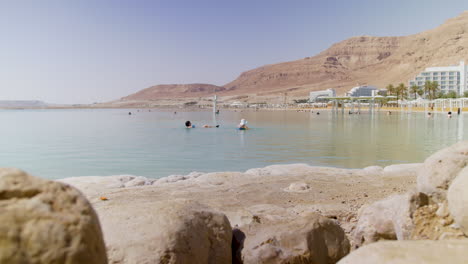 2-ältere-Touristen-Schwimmen-Im-Toten-Meer-–-Und-Genießen-Die-Morgensonne-–-Salzblöcke-Im-Vordergrund