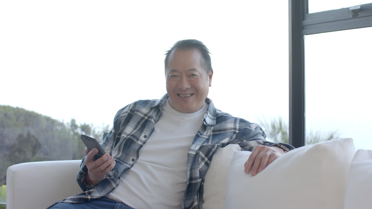 Smiling man holding smartphone sitting on couch in bright living room
