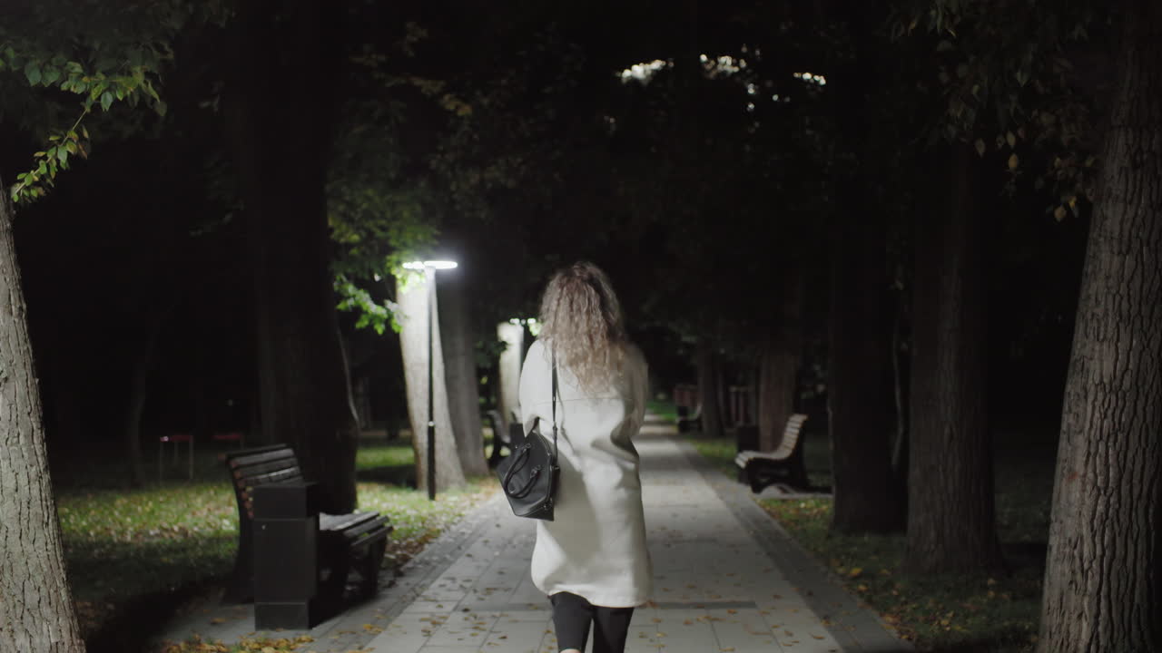 Back view of woman with curly hair in white coat walking alone on dimly lit park pathway at night, carrying black handbag, surrounded by benches, trees, fallen leaves and soft lamp illumination