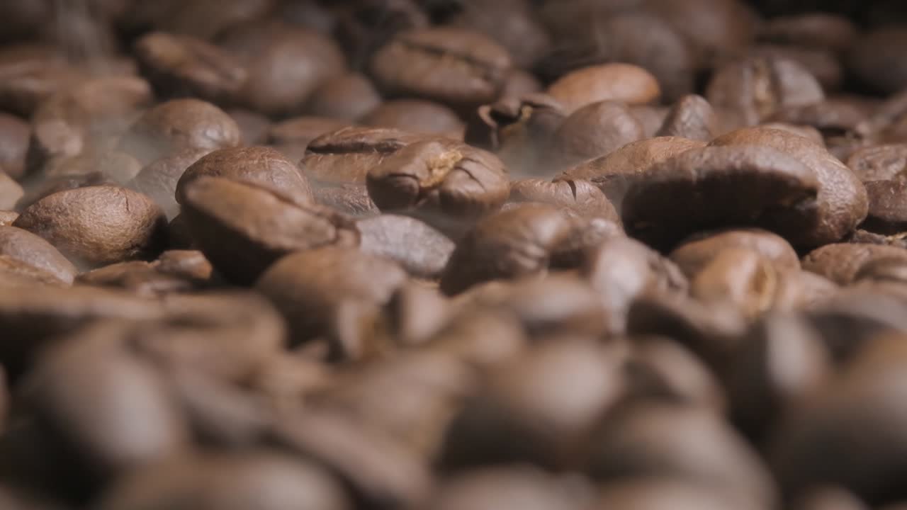 Close up slow motion shot of beans falling onto a full tray of roasting coffee beans, the impact disturbing the aromatic smoke being released as the beans bounce and come to rest