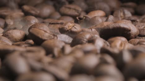 Close-up-slow-motion-shot-of-beans-falling-onto-a-full-tray-of-roasting-coffee-beans,-the-impact-disturbing-the-aromatic-smoke-being-released-as-the-beans-bounce-and-come-to-rest