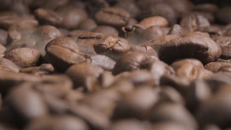 close up slow motion shot of beans falling onto a full tray of roasting coffee beans, the impact disturbing the aromatic smoke being released as the beans bounce and come to rest