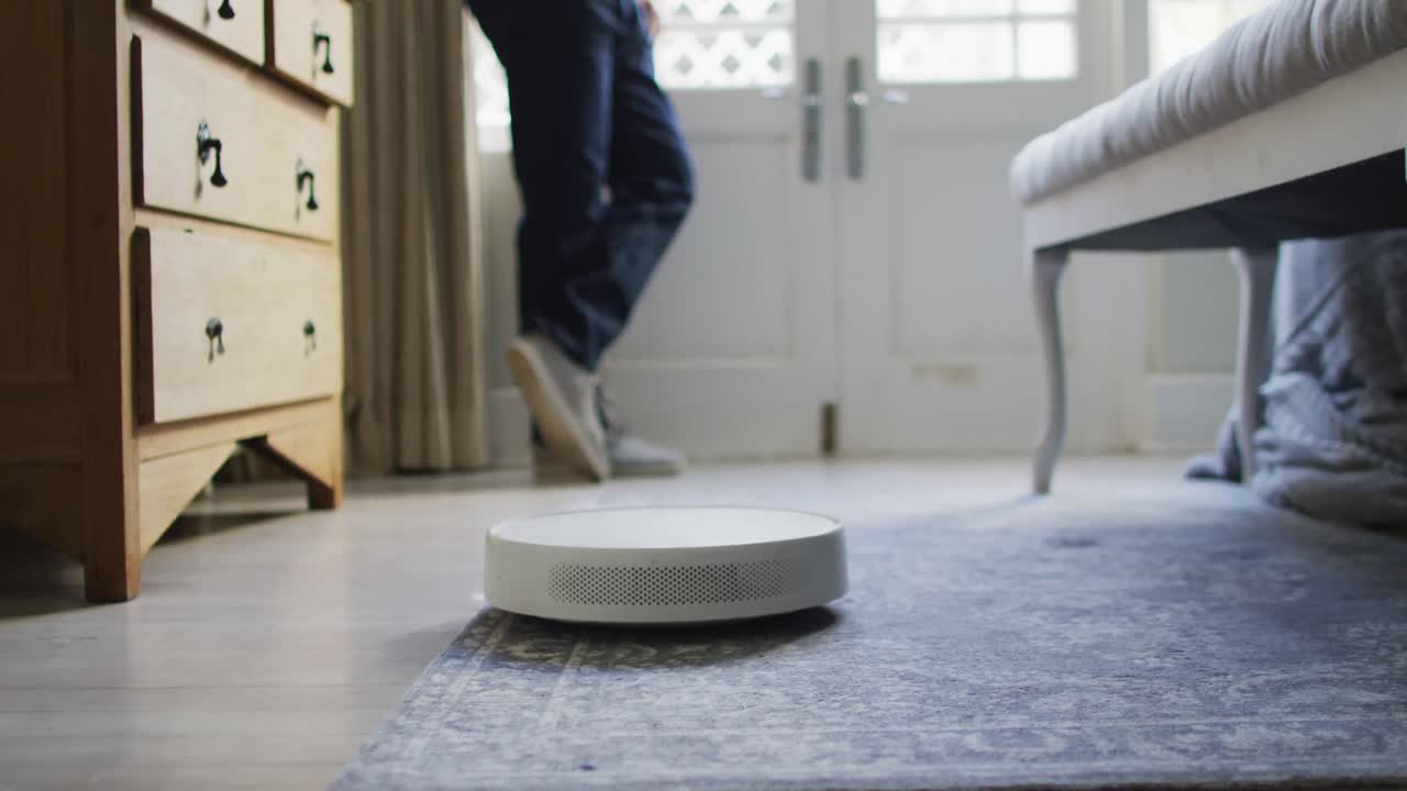 Midsection of caucasian man looking through window with robot vacuum cleaner on floor in bedroom