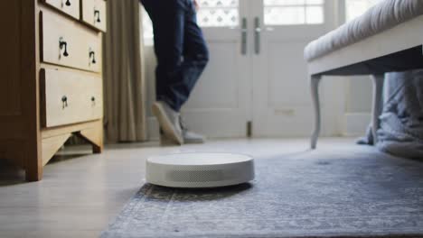 Midsection-of-caucasian-man-looking-through-window-with-robot-vacuum-cleaner-on-floor-in-bedroom