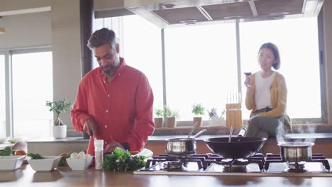 Happy-diverse-couple-cooking-together,-chopping-vegetables-and-drinking-wine-in-kitchen