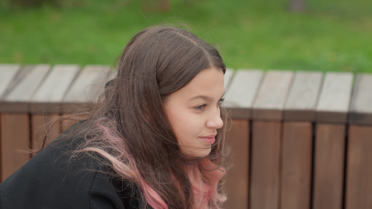 studente caucasico seduto su una panchina di legno, capelli tinti di rosa sulle spalle, che guarda in basso e poi di lato, parco sullo sfondo con erba verde, tonalità autunnali tenui, felpa con cappuccio e abbigliamento casual, pensieroso