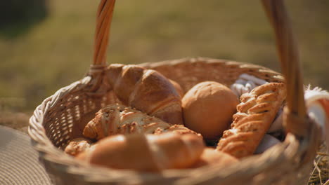 close-up of fresh golden croissants and bread rolls in woven picnic basket with soft cloth, placed on hay with woven hat nearby, sunlit countryside field in blurred background