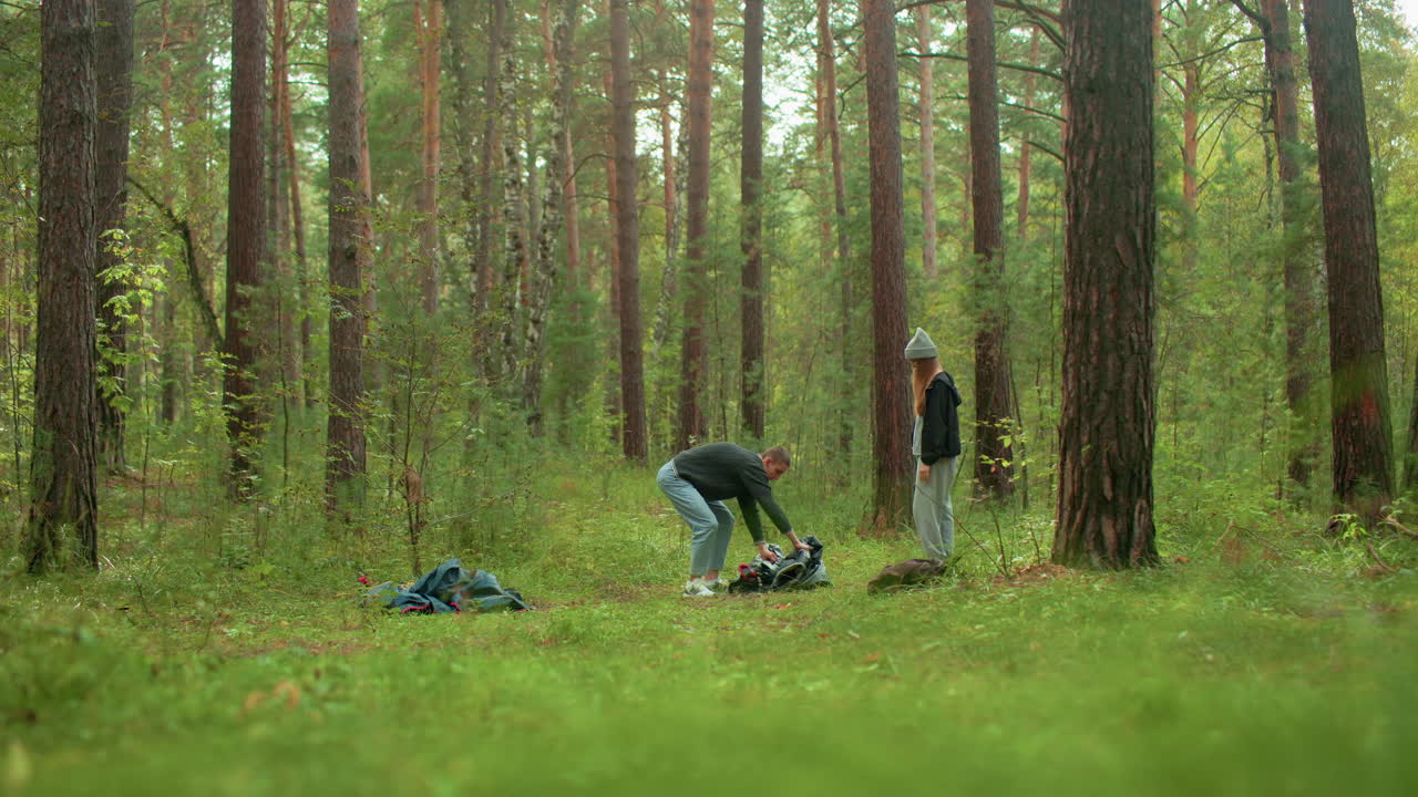 young man bends over unpacking tent while young woman watches in quiet forest clearing, surrounded by tall pine trees, scattered gear and camping supplies, during outdoor adventure in nature