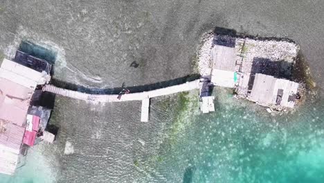 Aerial-top-view-Man-jump-on-old-wooden-pier-from-palafito-stilt-house,-Los-Roques-Venezuela