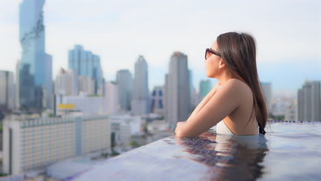 Mientras-Se-Apoya-En-El-Borde-De-La-Piscina-Infinita-De-Un-Resort-En-La-Azotea,-Una-Mujer-Atractiva-Mira-El-Horizonte-De-La-Ciudad-De-Bangkok,-Tailandia