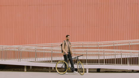 young american man in formal clothes looking at the camera while standing with his bike in front of a prefab metal building