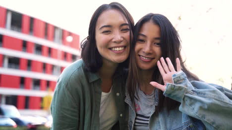 Two-Happy-Japanese-Female-Friends-Waving-At-Camera-And-Sending-Air-Kiss-While-Laughing-And-Enjoying-Time-Together-Outdoors