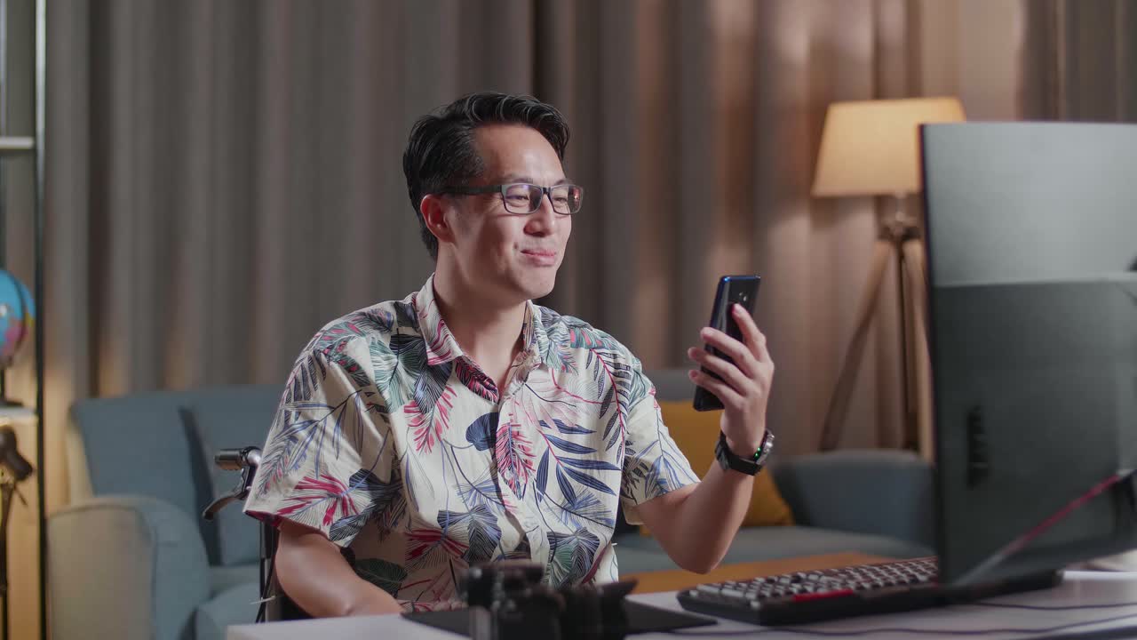 Asian Man In Wheelchair Waving Hand And Having A Video Call On Smartphone While Editing The Video By A Desktop Next To The Camera At Home