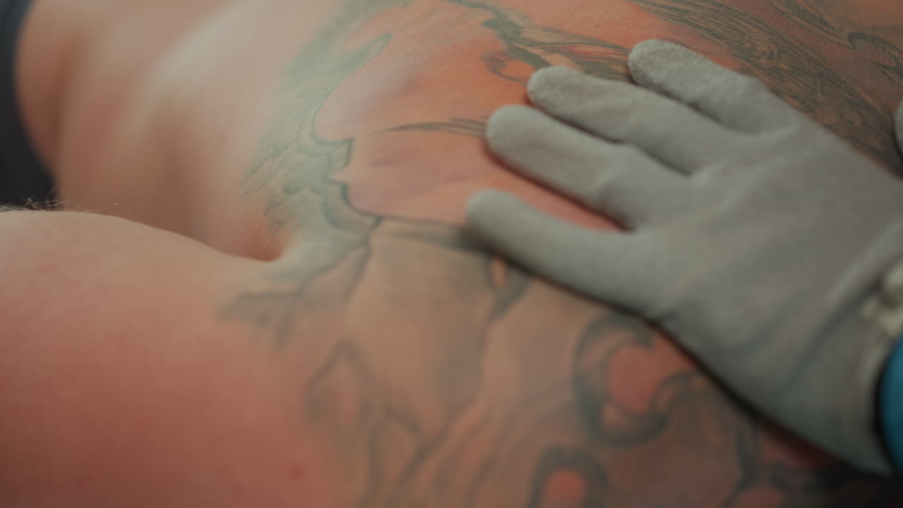 primo piano della schiena tatuata di un uomo caucasico, estetista che esegue un massaggio a microcorrente con guanto applicando una leggera pressione conduttiva sopra un tatuaggio nella parte bassa della schiena in un ambiente clinico con luci soffuse, catturando la texture della pelle e il dettaglio dell'inchiostro