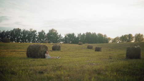 woman seated on ground, resting back against large hay bale in open farmland, peaceful rural landscape with scattered hay bales, warm evening light, and serene countryside setting