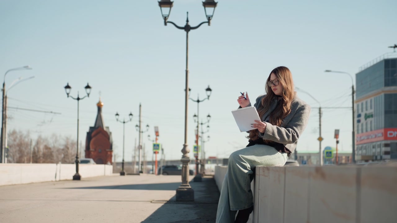 Pretty caucasian student reading papers outdoors, concrete ledge by city lamp, soft sunlight, wool jacket and jeans, notebook and pen in lap, thoughtful expression, distant church tower, calm