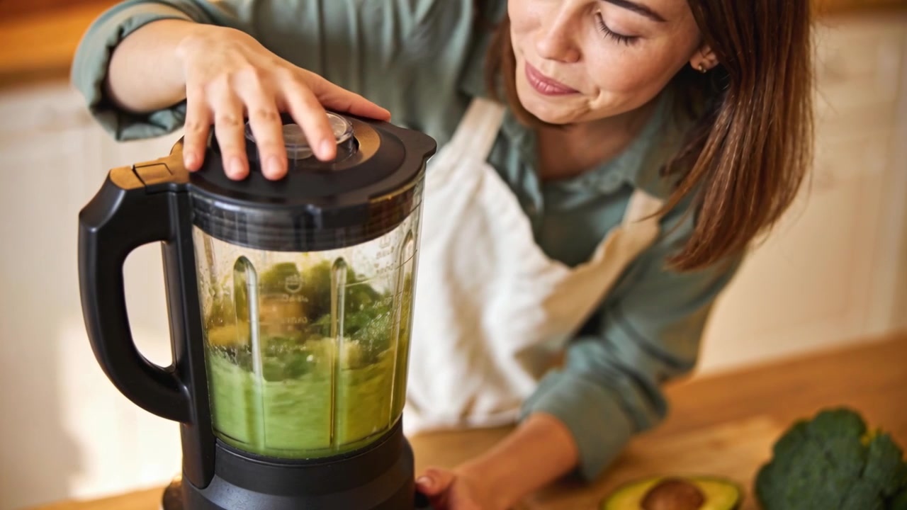 Mulher de avental colocando kiwi e verduras, verificando a tampa do liquidificador, batendo na cozinha para fazer uma bebida