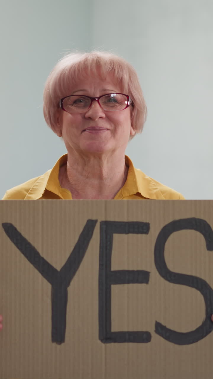 elderly woman picks up poster with inscription YES sitting on couch in the room of house, smiling, wearing glasses, looks good in yellow shirt and jeans, agrees