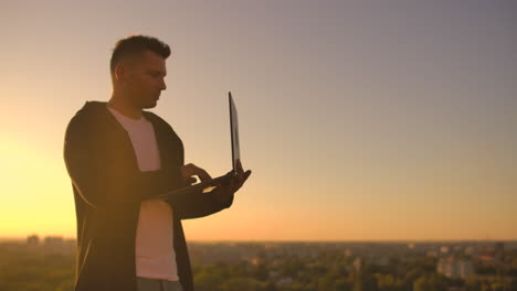 usando un portátil en el techo con vista a la ciudad y gráfico de divisas. concepto de hacking y estadísticas. un hombre al atardecer en cámara lenta escribiendo código de software en un portátil hacker