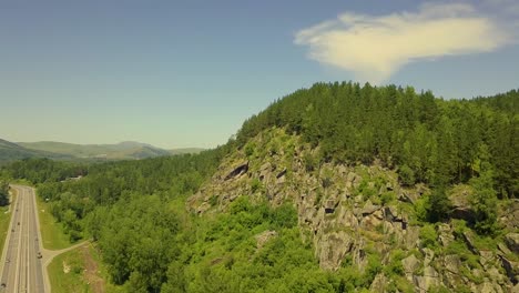 Aerial-Flying-along-stone-mountain-with-a-coniferous-forest-Below---the-highway-driving-cars