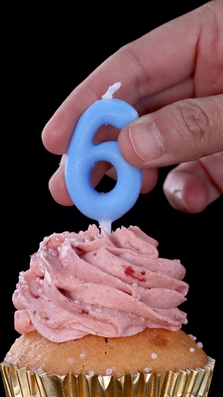 Una mano inserta una vela de cumpleaños azul con el número seis en un cupcake glaseado de color rosa