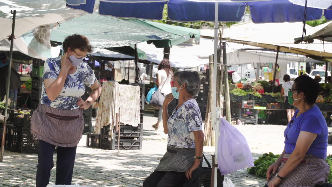 Medium shot of three farmers talking, while wearing masks and selling their vegetables in a fair, after the end of lockdown (deconfinement), in Barcelos, Portugal.